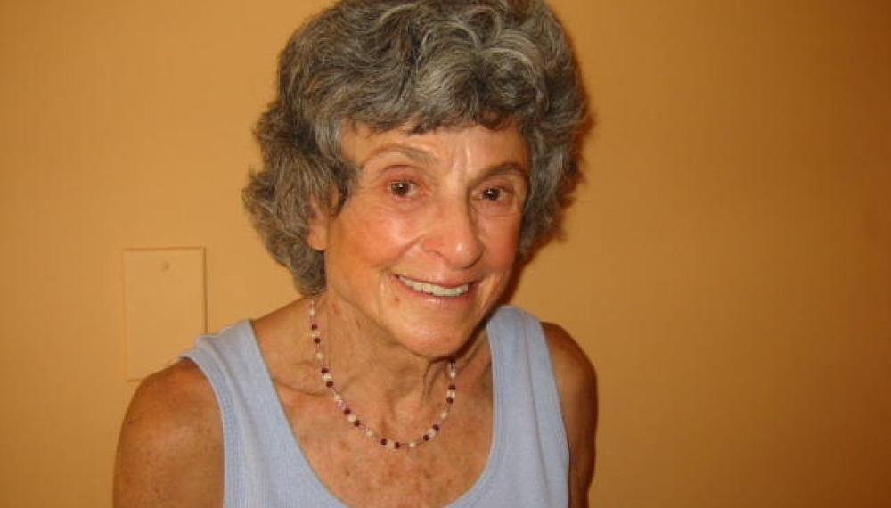 Alice Kahn Ladas stands in front of a yellow wall wearing a blue tank top. 