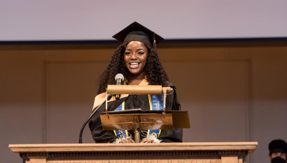 Beth Nanjala Luvisia wears a cap and gown as she stands behind the podium delivering a speech during Commencement 2022.