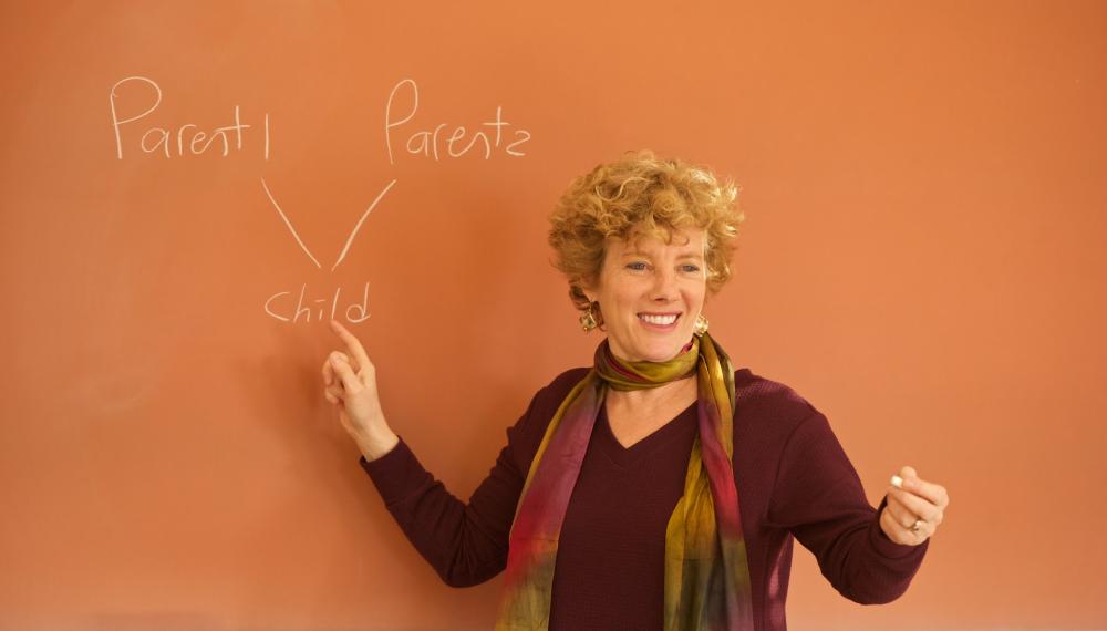 Marsha Kline Pruett stands in front of a blackboard with a diagram reading parent 1, parent 2, child drawn on it.