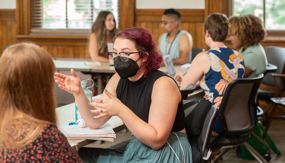 Two students sit in a classroom engaged in discuss. One has their back to the camera. The student facing the camera is wearing a mask and gesturing with both hands.