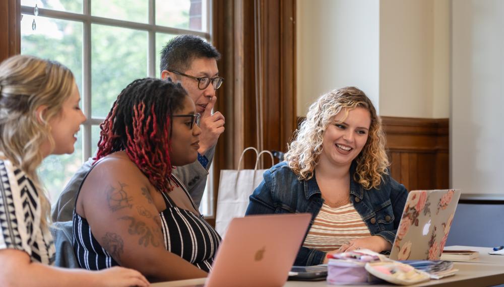 Three students sit in a classroom with laptops while a faculty member stands nearby engaged in conversation. 
