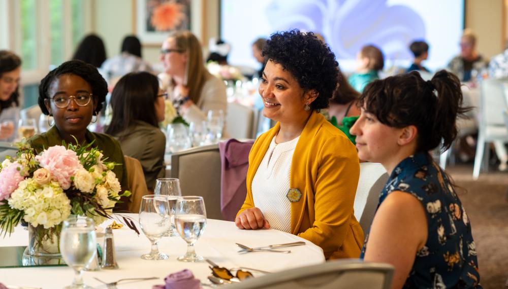 Three people sitting at a table with white tablecloth and flowers. Behind are more people sitting with a screen in the far background