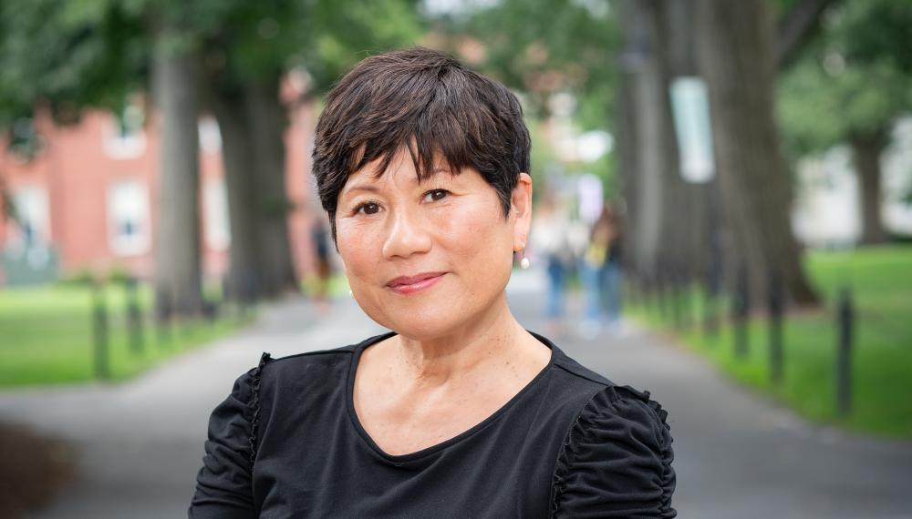 Marianne Yoshioka stands behind a granite post, leaning on it smiling with green trees behind