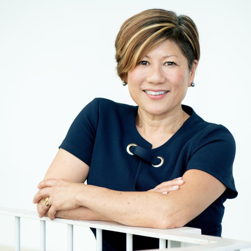 Dean Marianne Yoshioka leans over a railing in the campus center. She is wearing a blue dress.