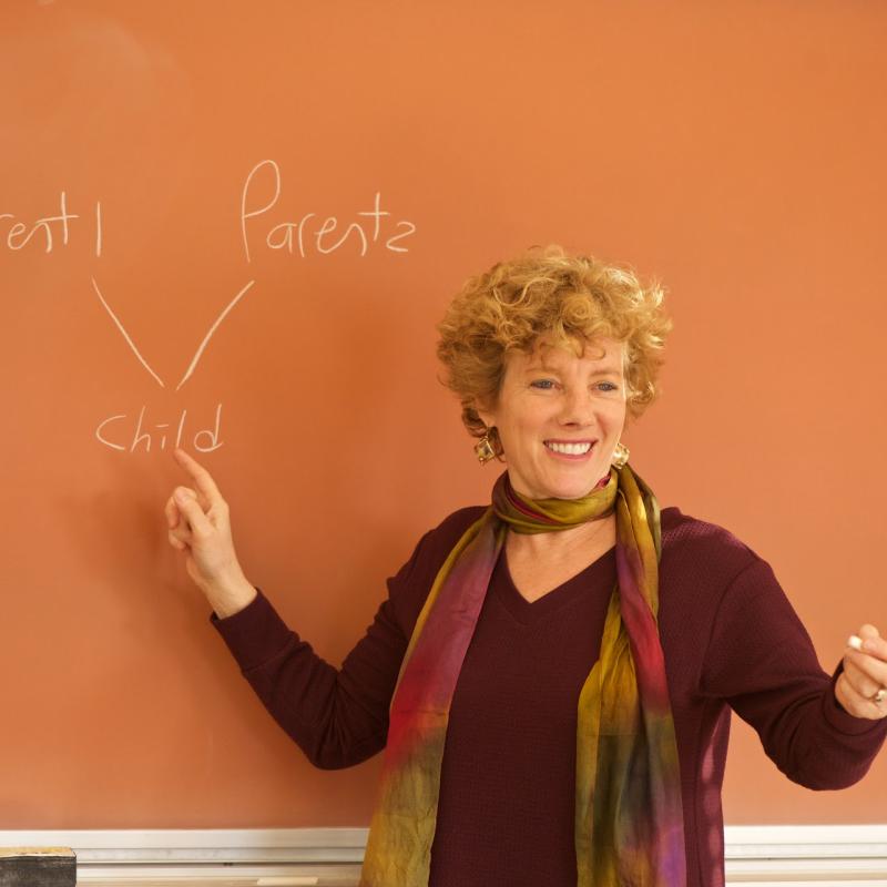 Marsha Kline Pruett stands in front of a blackboard with a diagram reading parent 1, parent 2, child drawn on it.