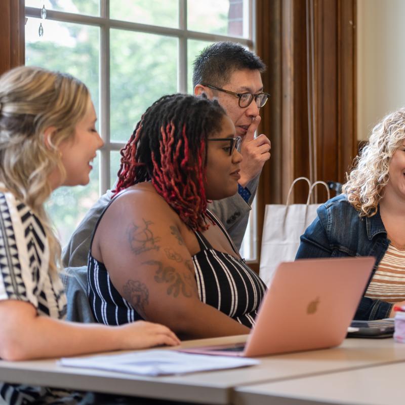 Three students sit in a classroom with laptops while a faculty member stands nearby engaged in conversation. 