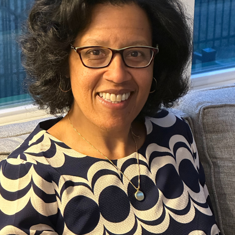 Jennifer Ashby Bullock headshot, sitting on grey couch wearing black glasses and black and white shirt