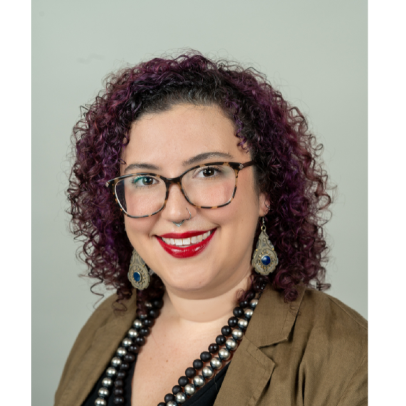 Hannah Biskind wearing glasses and a tan blazer, earrings and necklaces against a white wall