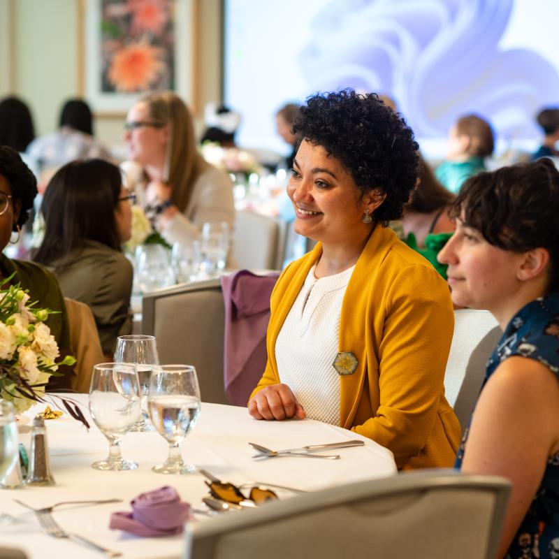Three people sitting at a table with white tablecloth and flowers. Behind are more people sitting with a screen in the far background