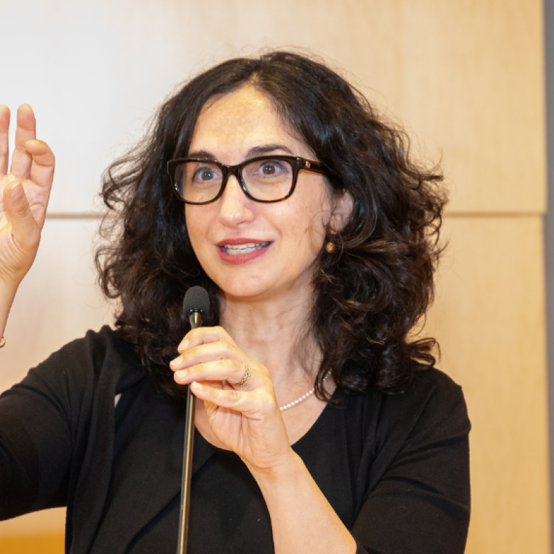 Ora Nakash speaking holding a mic with wood paneling behind