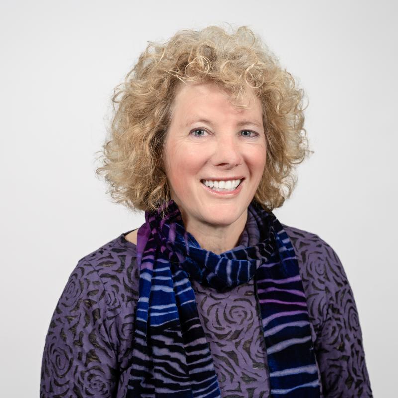 Marsha Kline Pruett wearing purple shirt and blue striped scarf smiling with white background