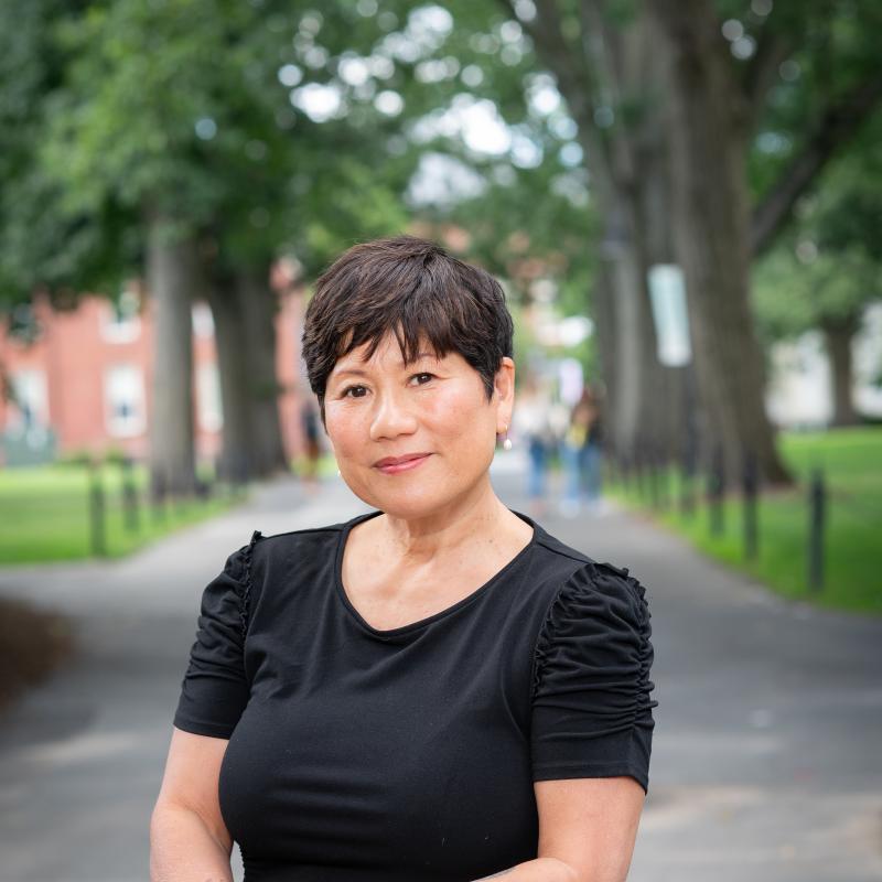 Marianne Yoshioka stands behind a granite post, leaning on it smiling with green trees behind