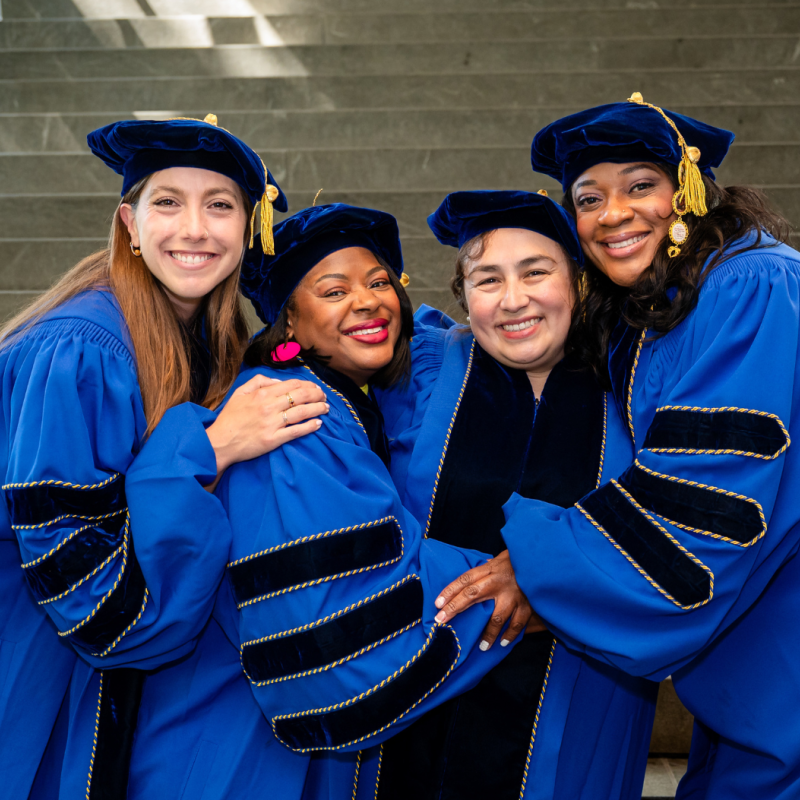 Four phd graduates in blue academic robes and tams hug and smile in front of cement stairs