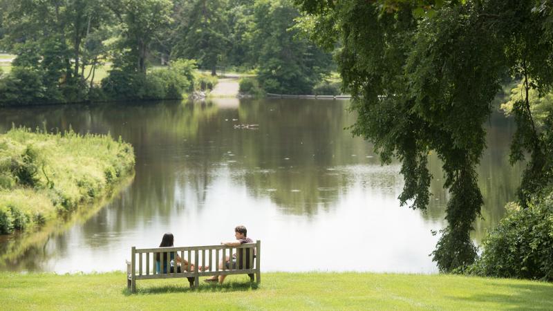 Photo of two students sitting on a wooden bench overlooking Paradise Pond at Smith College. 