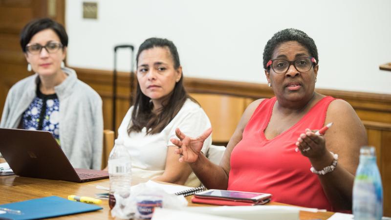 Three continuing education participants sit at a wooden table with note taking supplies in front of them, one of the participants in a red tank top gestures with their pen as they talk towards the front of the room.