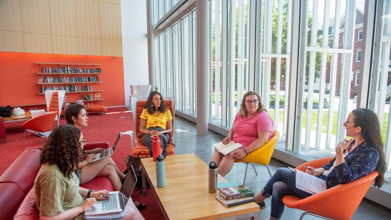 Five students sit around a coffee table in the Campus Center. There is a bright orange wall in the background and floor to ceiling windows along one side. 