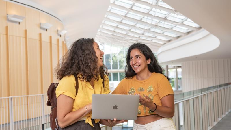 Two students stand on the second floor of the Campus Center. One holds a laptop. They are looking at one another and laughing. 