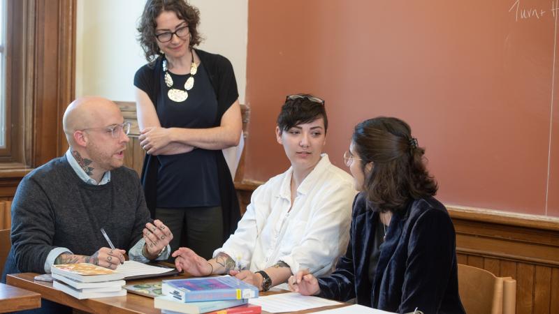 A professor stands next to three students who are seated at a table. The professor is listening to the students who are engaged in conversation. There are books and papers all over the table. 