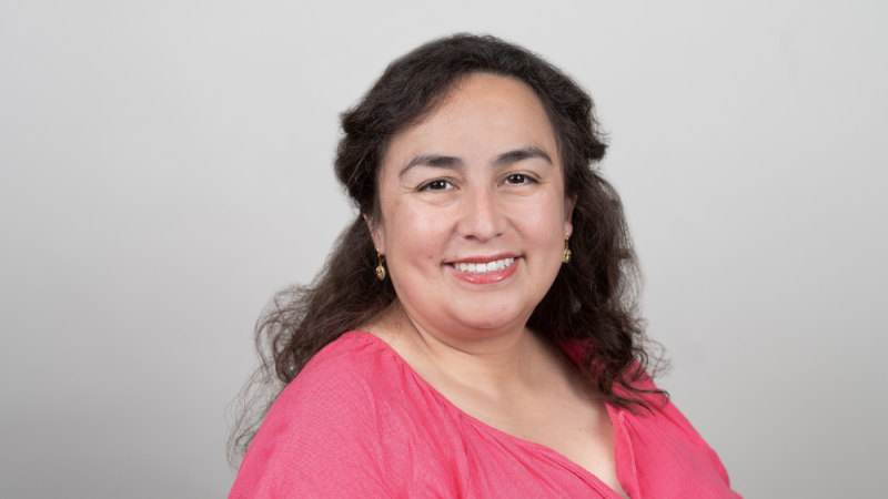 Maria Maldonado-Morales wearing bright pink shirt against grey wall smiling